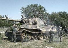 two men standing next to a tank on top of a grass covered field with trees in the background