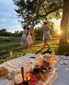 three women dancing around a table with food and drinks on it in the evening sun