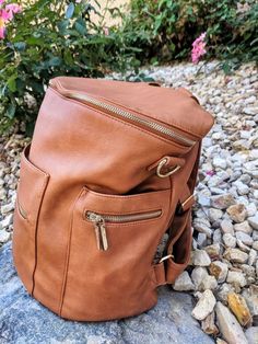 a brown leather backpack sitting on top of a rock next to some rocks and flowers