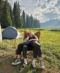 two people are sitting on a chair in the grass near a tent and campfire