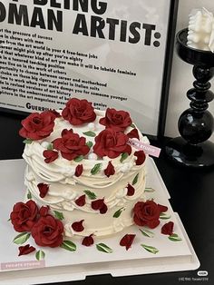 a white cake with red roses on it sitting next to a black vase and sign