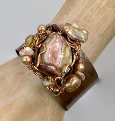 "Pink and shades of soft browns and beige....  Starting with a copper cuff bracelet that I have hammered and heated for an amazing color... I then chose the large jasper stone that is the centerpiece of this bracelet. I love the swirling shades of pink and cream.  I encased it in a freeform, forged copper bezel with a unique organic shape to it.  I have given it an artistic and organic wire wrap . For additional accents I have used beads in stone, glass and pearls to enhance the shades of the st Elegant Brown Cuff Bracelet As Gift, Elegant Brown Cuff Bracelet Gift, Adjustable Copper Artisan Cuff Bracelet, Artisan Adjustable Copper Cuff Bracelet, Adjustable Artisan Copper Cuff Bracelet, Artisan Copper Cuff Bracelet Adjustable, Artisan Copper Cuff Bracelet With Adjustable Fit, Bronze Hand Wrapped Cuff Bracelet Gift, Bronze Hand Wrapped Cuff Bracelet For Gift