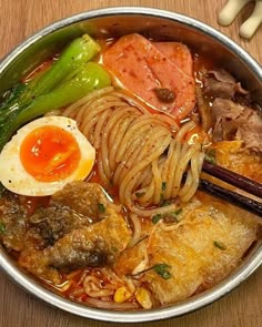 a metal bowl filled with meat, noodles and vegetables next to chopsticks on a wooden table