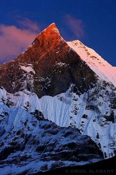 the top of a mountain covered in snow at sunset with clouds and blue sky behind it