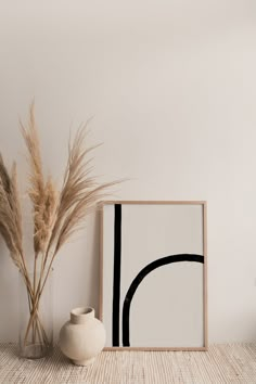 a white vase sitting next to a painting on top of a wooden table with a plant in front of it