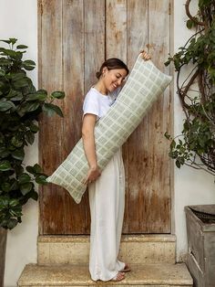 a woman holding a large pillow in front of a wooden door with potted plants