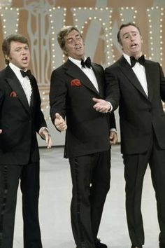 three men in tuxedos stand on stage with their hands out to the side