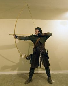 a man dressed in medieval clothing holding a bow and arrow while standing next to a wall