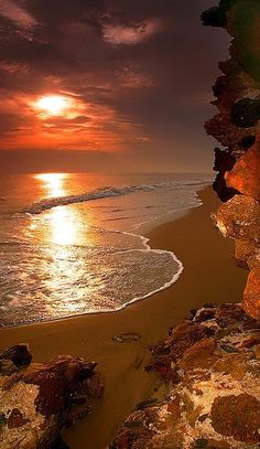 the sun is setting over the water at the beach with rocks in front of it