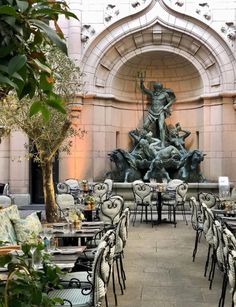 an outdoor dining area with tables, chairs and a statue in the center surrounded by greenery