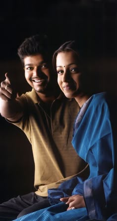 a man and woman sitting next to each other in front of a black background with one pointing at the camera