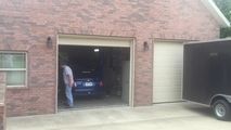 a man standing in the doorway of a garage next to a blue car and a black trailer