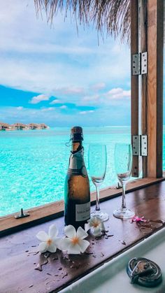 a bottle of wine and two glasses on a table by the water with flowers in front of it