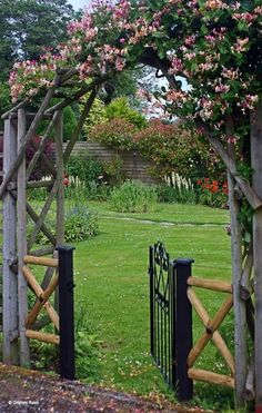 an open gate in the middle of a lush green yard with pink flowers on it