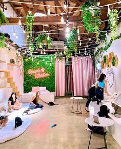 several people sitting on couches in a room with plants hanging from the ceiling and pink curtains