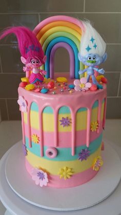 there is a birthday cake with pink frosting and rainbow decorations on it, as well as two little trolls