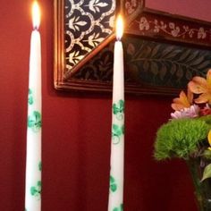 two white candles sitting on top of a table with flowers in the vase next to it