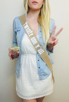 a woman with blonde hair wearing a sash and making the peace sign while holding a drink in her hand