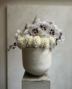 a white vase filled with lots of flowers on top of a cement block in front of a wall