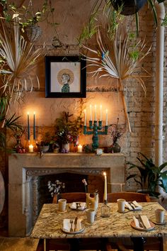 a dining room table with candles and plants on the fireplace mantel area in front of it