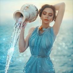 a woman in a blue dress holding a water pitcher over her head while standing next to the ocean