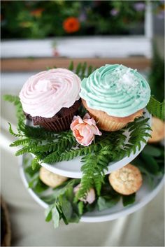 three cupcakes with frosting and greenery on a cake stand in front of flowers