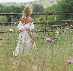 a woman in a white dress is walking through the grass with flowers behind her back to the camera