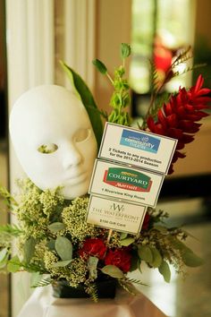 a white mask sitting on top of a vase filled with flowers and greenery next to a sign