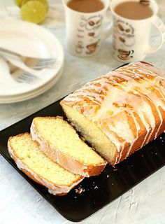 a loaf of lemon pound cake sitting on top of a black platter next to two cups of coffee