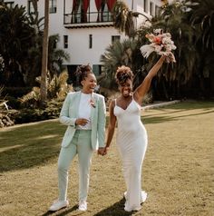 two women in white outfits holding flowers and walking through the grass with palm trees behind them