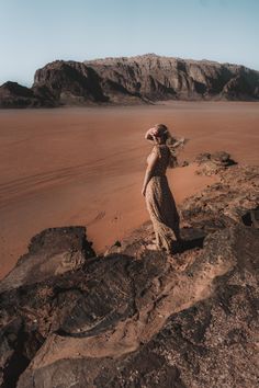 a woman standing on top of a rocky hill next to a sandy desert area with mountains in the background