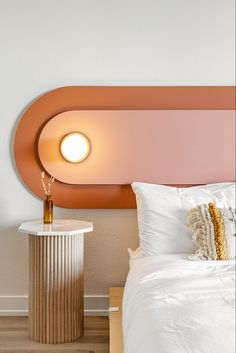 a white bed sitting under a round light on top of a wooden table next to a night stand