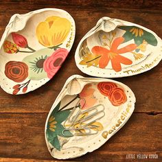 three decorative trays with flowers painted on them sitting on a wooden table, one is holding a paper clip and the other has scissors in it