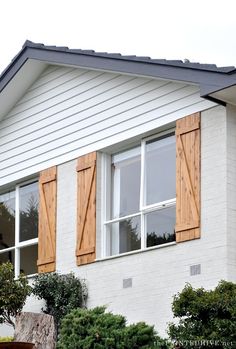 a white house with wooden shutters and green bushes in front of the window area