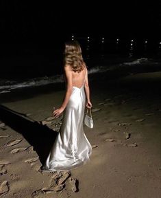 a woman in a white dress walking on the beach at night with her back to the camera