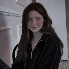 a woman with long brown hair wearing a black shirt and smiling at the camera while standing in front of a white wall