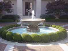 a fountain in front of a building with trees and bushes around it's perimeter