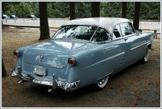 an old blue car parked in front of some trees and dirt with no leaves on it