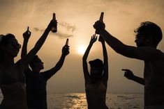 group of people raising their hands in the air at sunset on the beach with beer bottles