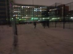 two people are walking in the snow near a fence at night with buildings in the background