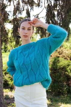 a woman standing in front of a tree wearing a blue sweater and white skirt with her hands on her head