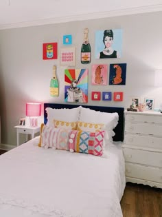 a white bed topped with lots of pillows next to a wall covered in pictures and paintings