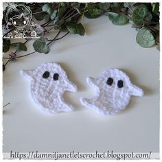 two crocheted ghost faces sitting on top of a table next to green leaves