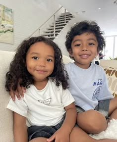 two young children sitting on a couch in a living room with white walls and stairs