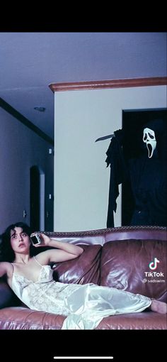 a woman laying on top of a brown couch in front of a black ghost hanging from the wall