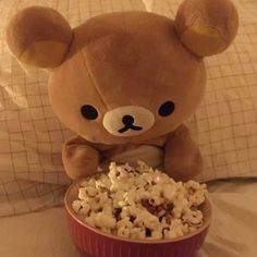 a brown teddy bear sitting on top of a bed next to a bowl of popcorn