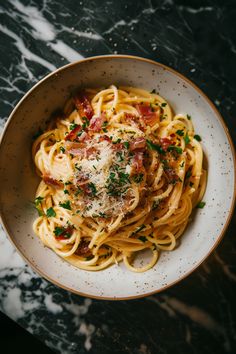 a white bowl filled with pasta covered in bacon and parmesan on top of a marble table