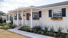 a white house with black shutters and flowers on the front porch, along side a sidewalk