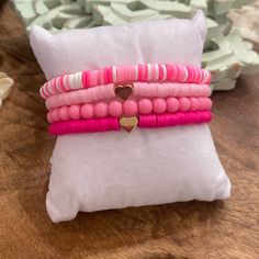 three pink and white bracelets with a heart on them sitting on a pillow next to a plant