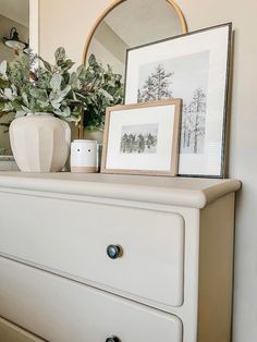 a white dresser topped with a vase filled with flowers next to a mirror and framed pictures